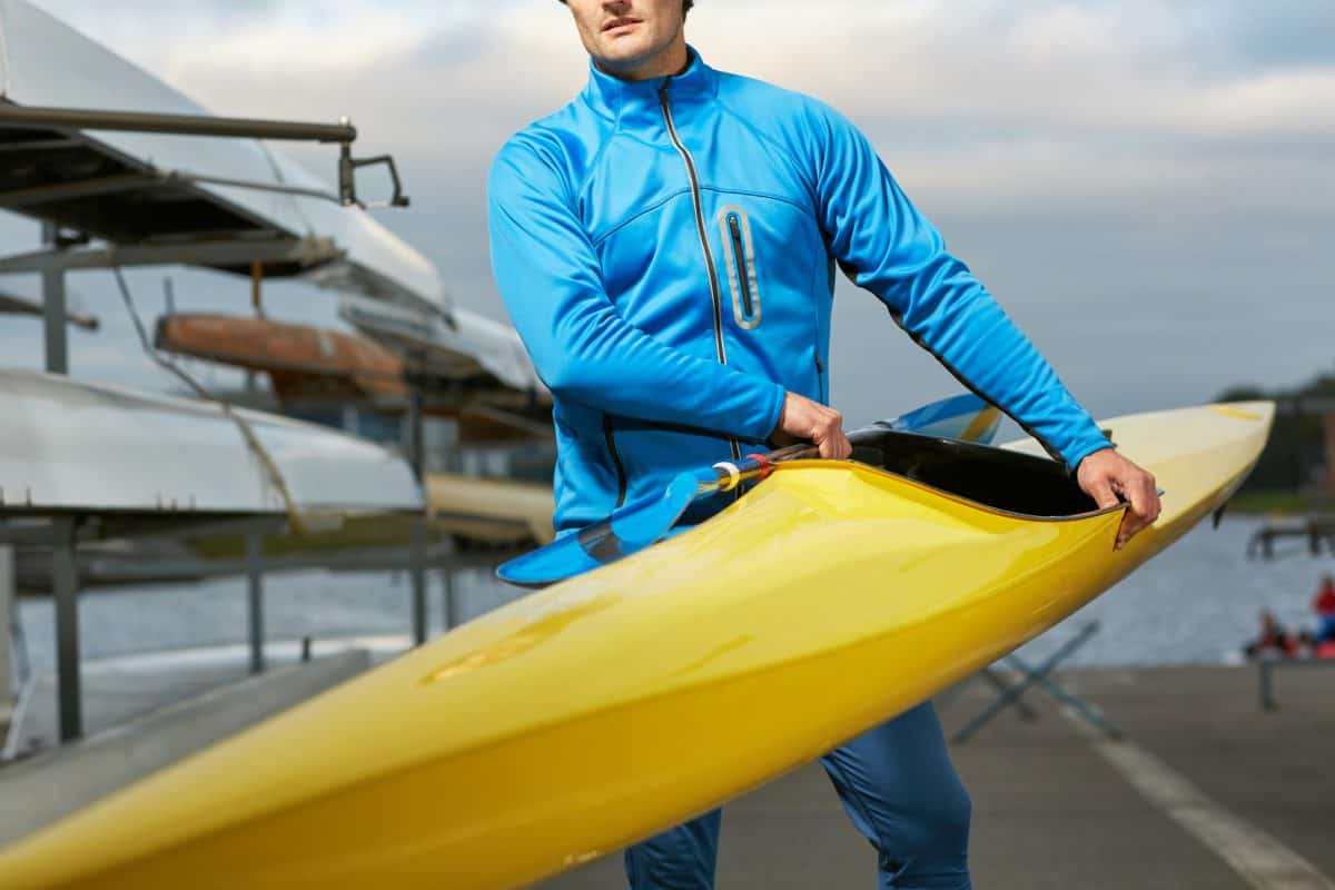 Male athlete in swimming sportswear carrying yellow kayak and paddle outdoors after training on the river,