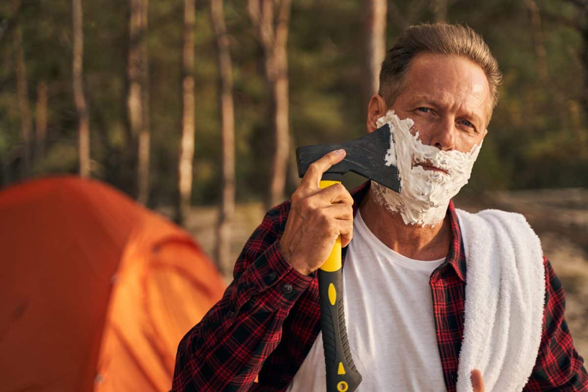 Mature man shaving with axe while going camping